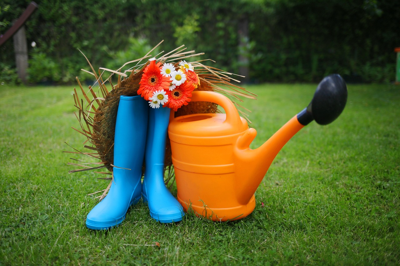 arrosoir en plastique orange, bottes en caoutchouc bleues et chapeau de paille avec des fleurs posés dans l'herbe